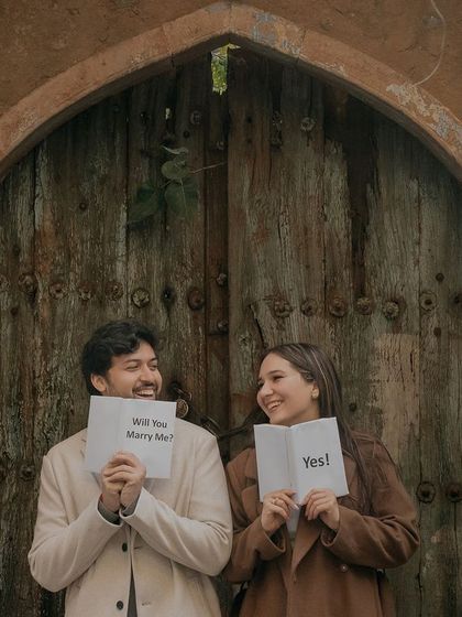 A playful and charming pre-wedding moment in Old Delhi. The "Will You Marry Me?" signs add a sweet, personal touch, while the rustic door provides a wonderfully textured and authentic backdrop.
