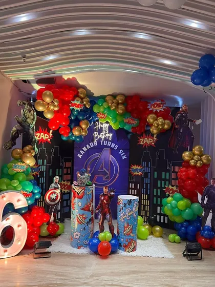A full view of our Avengers-themed birthday party setup for a six-year-old. The decoration includes a city skyline backdrop, character standees of superheroes like Iron Man and Captain America, a large light-up number '6', and a vibrant balloon garland.