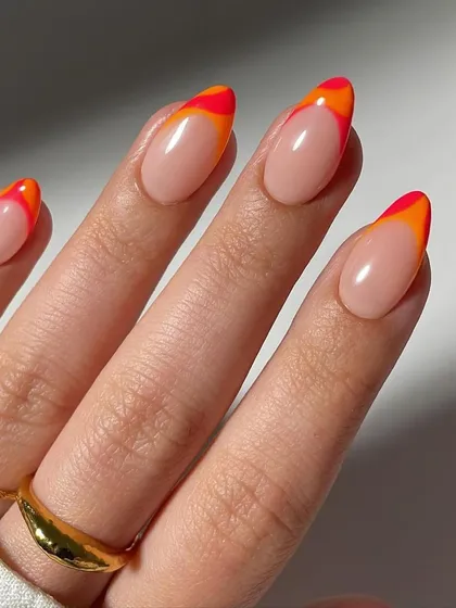 A close-up of a vibrant and fiery double French manicure. The almond-shaped nails have a clean nude base with layered red and orange tips, creating a beautiful sunset effect.