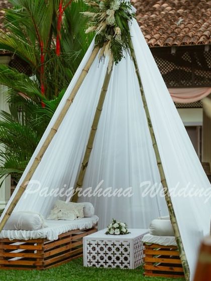 A cozy boho lounge area for a Mehendi. The setup includes a simple teepee tent, a wicker peacock chair, and seating made from wooden crates, creating a relaxed and stylish vibe.