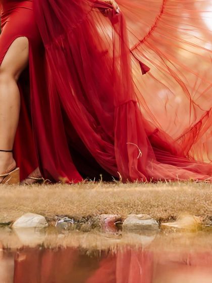 A close-up detail shot focusing on the flowing red fabric of a maternity gown against the natural grass and water's edge. This artistic photo highlights the texture and movement of the dress.