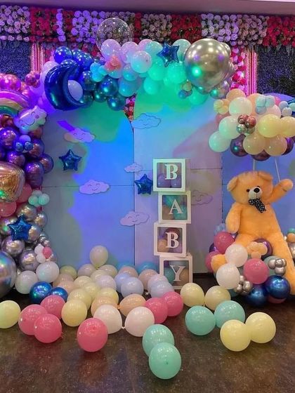 This colorful baby shower setup includes a large teddy bear prop sitting on a cloud of balloons. The backdrop features "BABY" display boxes, rainbow and moon foil balloons, and a floor scattered with pastel balloons.