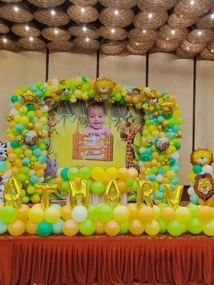 A grand stage setup for a jungle-themed first birthday. The decoration features a large backdrop with a personalized photo and name, surrounded by a lush balloon arrangement.
