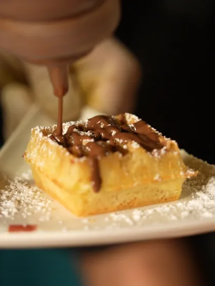 A freshly made waffle square being generously drizzled with chocolate sauce. This action shot from our live waffle station captures a moment of pure indulgence.