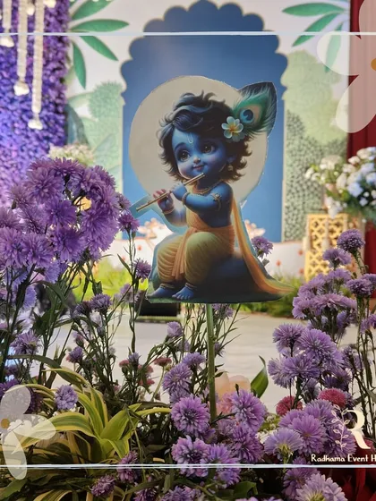 A close-up of a "Little Krishna" themed naming ceremony. A cute cutout of Lord Krishna playing the flute is nestled among purple flowers, adding a divine and playful touch to the overall decor.