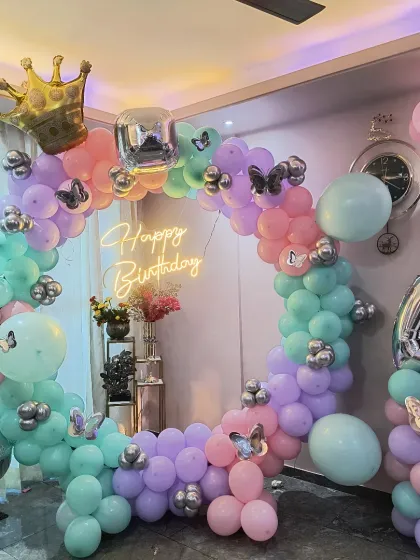 A magical princess-themed birthday party decoration. This features a full circular balloon arch in pastel shades of pink, purple, and mint green, accented with silver butterflies, a golden crown, and a 'Happy Birthday' neon sign.
