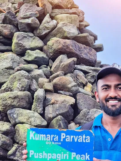 A trekker proudly holding the signboard at the summit of Kumara Parvatha, also known as Pushpagiri Peak.