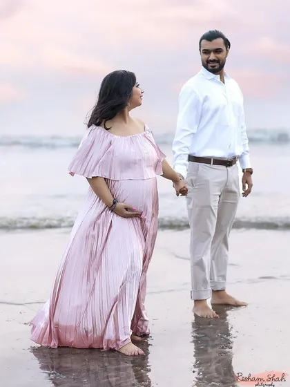 A beautiful wide shot of the couple walking barefoot along the shoreline. The mom-to-be's pink dress flows gently in the breeze, creating a sense of peace and movement.