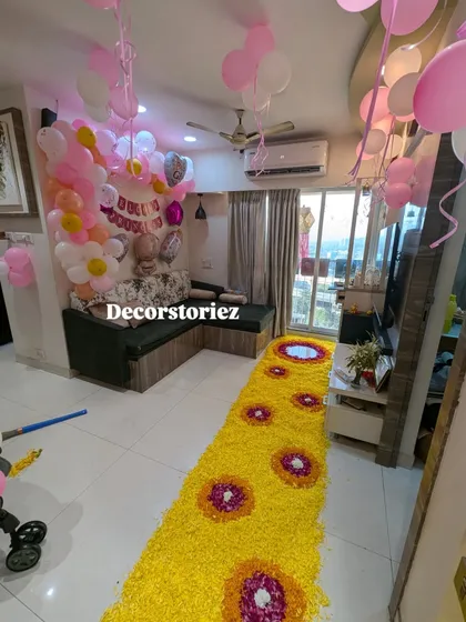 A complete room transformation for a baby girl's welcome. This setup combines a pink and white balloon arrangement in the background with a long, beautiful floral rangoli runner made of yellow and red flower petals.