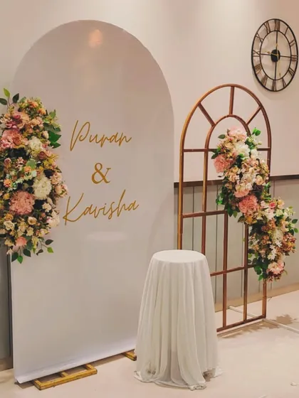 A clean and romantic engagement backdrop. I paired a white arched panel with the couple's names, "Puran & Kavisha," with a rustic window frame accent and soft floral decor.