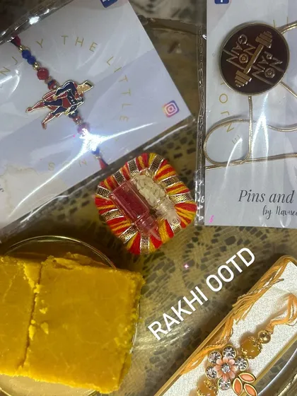 The perfect Rakhi OOTD (Outfit Of The Day) setup. This flat-lay shows our unique rakhis, including a Superman design and a "No Pain No Gain" lapel pin, ready for the celebration with traditional sweets.