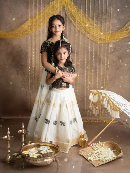 Two sisters dressed in beautiful pattu pavadai for our Onam-themed shoot. The setup includes a brass uruli with flowers and a traditional muthukuda umbrella.