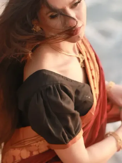 A candid shot capturing the wind in her hair by the sea. The off-shoulder blouse adds a modern touch to the traditional saree.