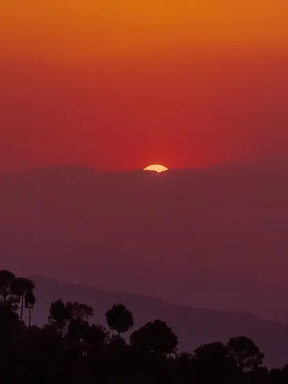 A vibrant red and purple sunset over the hills of Ranikhet. The sun is just a sliver on the horizon, casting a deep, warm glow over the entire landscape.