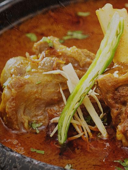 A close-up of a rich and hearty North Indian mutton curry. The tender meat on the bone is simmered in a flavorful gravy, garnished with fresh ginger and a green chili, representing the authentic taste of our 'The Charcoal Story' venture.