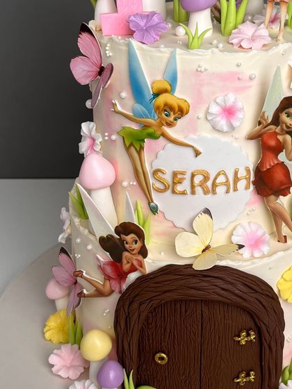 A close-up of our two-tiered fairy house cake, showcasing the hand-painted Tinkerbell figurine and other fairy friends. The details include sculpted mushrooms, butterflies, and a personalized nameplate in gold.