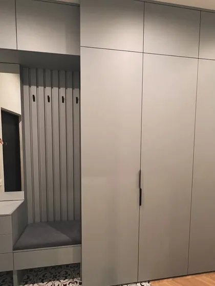 This modular wardrobe unit in Gurgaon includes a built-in entryway bench with a cushioned seat and coat hooks. The floor-to-ceiling design in a matte grey finish maximizes storage and adds a functional feature to the room.