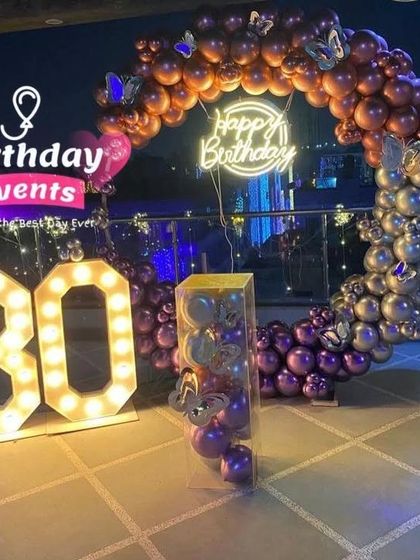 A glamorous outdoor terrace setup for a 30th birthday. The purple and rose gold balloon ring is accented with butterfly details and large marquee numbers.