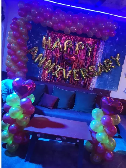 A cozy anniversary decoration in a dimly lit room. The setup includes a balloon arch, 'Happy Anniversary' foil letters, and heart-shaped foil balloons.