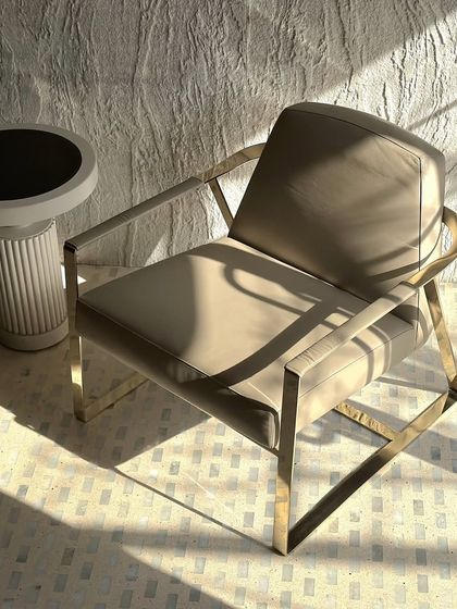 Another angle of the lounge chair, highlighting the clean lines of the gold frame and the smooth upholstery, set against a textured wall.