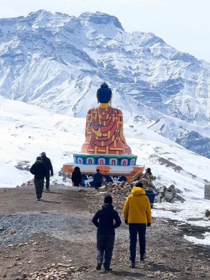 Pilgrims and travelers walking towards the Langza Buddha statue in Spiti, with snow-covered mountains behind.
