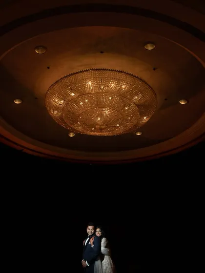 A dramatic portrait using negative space, with the couple positioned under a massive, ornate chandelier. The composition emphasizes the grandeur of the location and the intimacy of the couple.