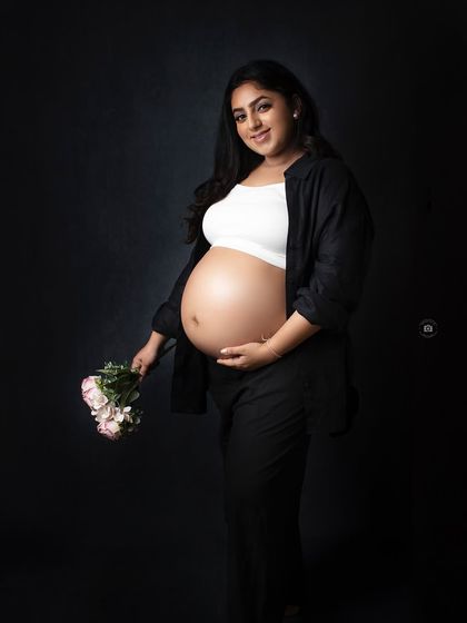A modern and confident solo portrait. The mother-to-be shows off her baby bump in a crop top and black pants against a dark background.