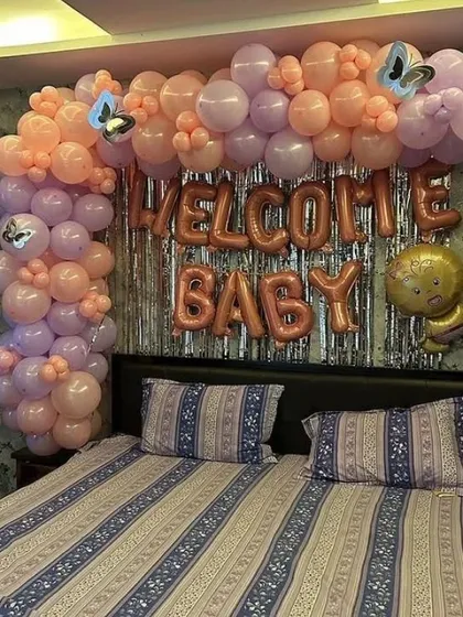 A lovely "Welcome Baby" decoration set up over a bed. The arch of pastel pink and lavender balloons, combined with a silver fringe curtain, creates a dreamy welcome for the newborn.