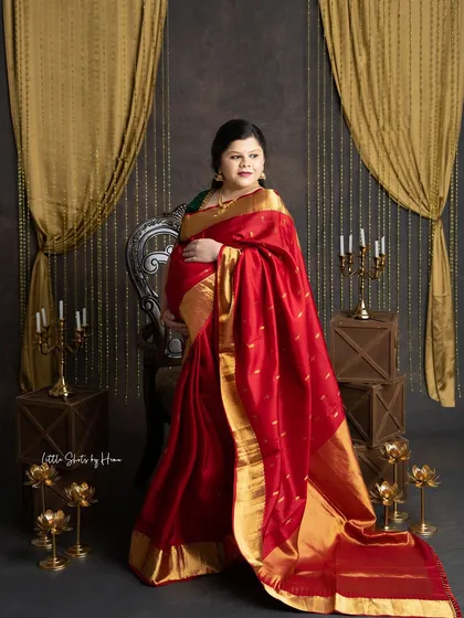 An elegant portrait of an expecting mother in a beautiful red silk saree with a wide gold border. She stands in a traditionally decorated studio with brass lamps and golden drapes.