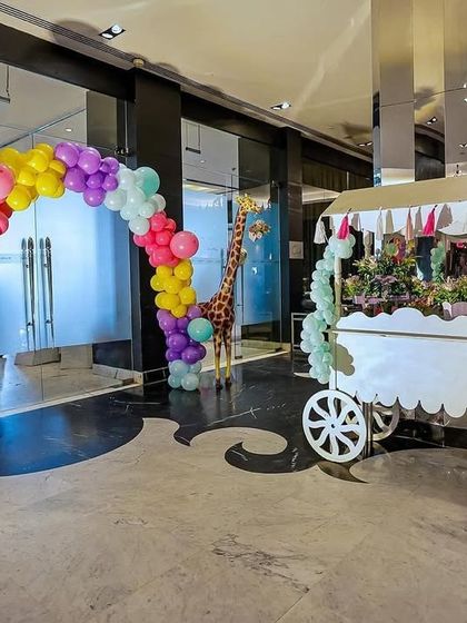 A full view of the vibrant indoor carnival entrance. The setup includes a colorful balloon arch, life-sized giraffe props, and a cart with a directional signpost for activities, games, and food.