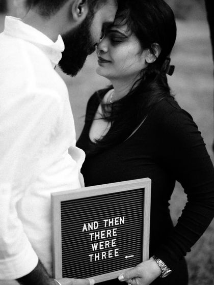 An intimate black and white close up. The couple is nose-to-nose, holding a letter board that reads "And Then There Were Three," a simple and powerful message.