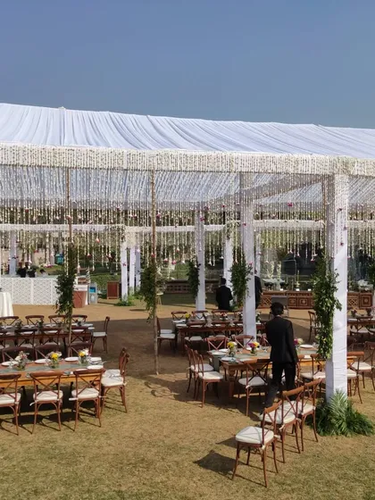 A beautiful and expansive daytime event setup under a large white tent. The decor features elegant guest seating with long wooden tables and hanging floral tassels, perfect for a sophisticated farmhouse lunch or brunch event.