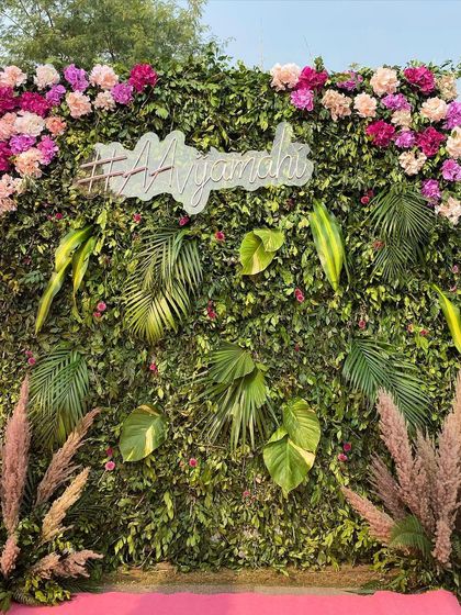 A lush green photo wall perfect for a tropical or garden-themed event. The wall is adorned with fresh monstera leaves and topped with a beautiful arrangement of pink and purple flowers, along with a custom neon hashtag sign.