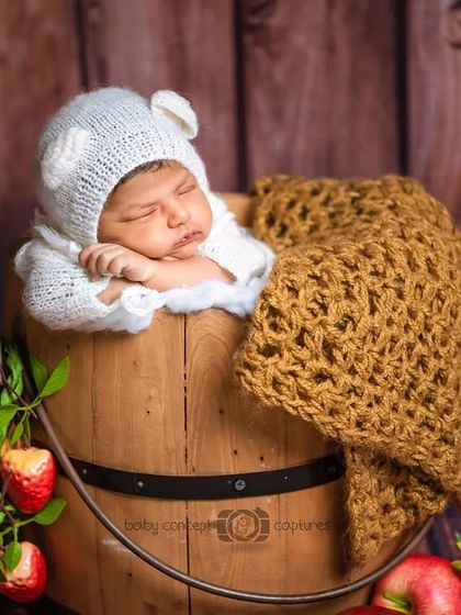 A cozy newborn wearing a knitted bear bonnet, sleeping soundly in a rustic wooden bucket. This setup uses natural textures and props for a warm, earthy feel.
