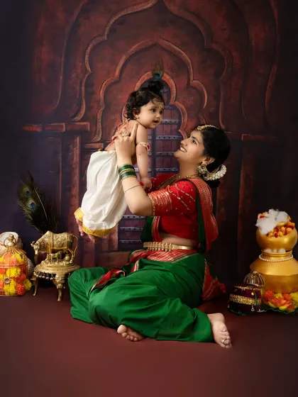 A heartwarming moment where a mother, dressed as Yashoda, lifts her baby Krishna into the air. The backdrop depicts a traditional Indian archway, adding to the classic feel.