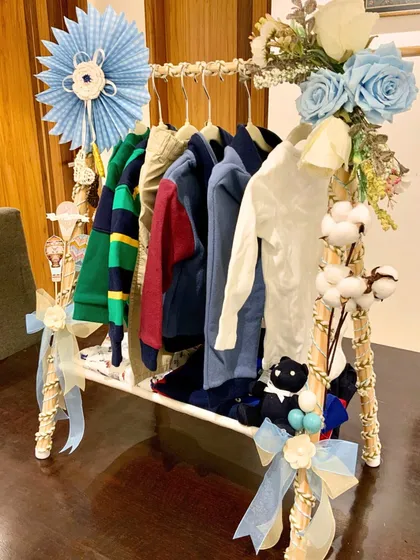 A unique and creative presentation for a baby boy's gift hamper. We have arranged a selection of high-quality baby clothes on a miniature decorative clothing rack, adorned with blue floral accents and delicate ribbons.