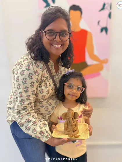 A mother and daughter duo share a happy moment, showcasing the beautiful clay Ganesha they created together in our family-friendly workshop.