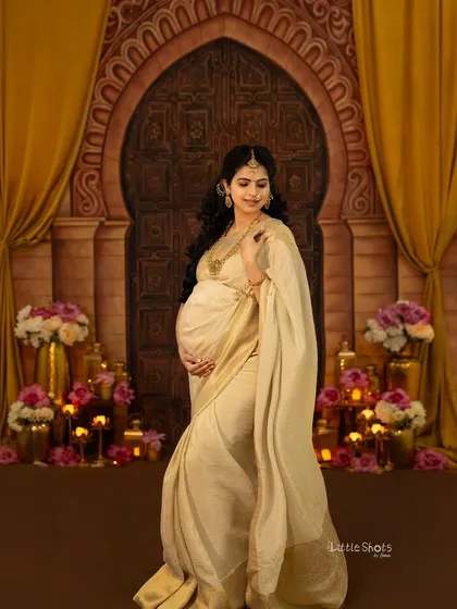 A stunning portrait of a mother-to-be in a cream and gold saree. She stands in a grand, palace-like setting with an arched doorway and floral decorations, looking like royalty.