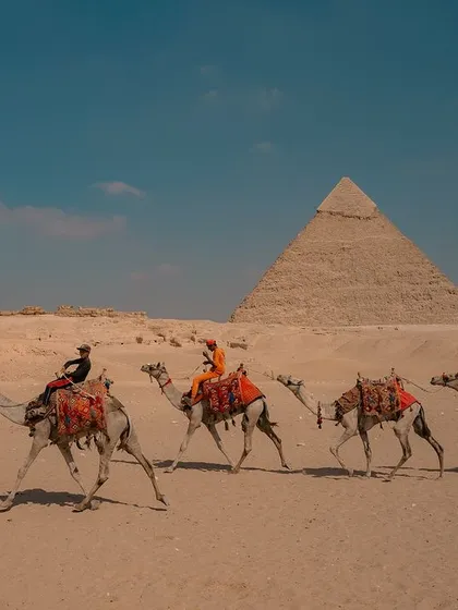 A caravan of camels carries tourists across the desert with the Great Pyramids of Giza in the background. A timeless scene of travel and exploration.