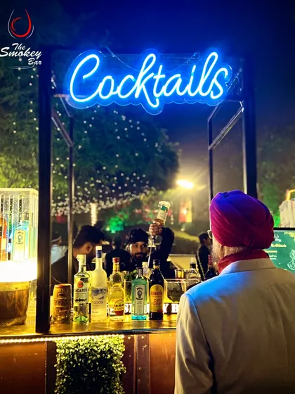 A guest enjoying the view of a bustling cocktail bar. The bright blue 'Cocktails' neon sign acts as a beacon, drawing people to the heart of the party where my mixologists are at work.