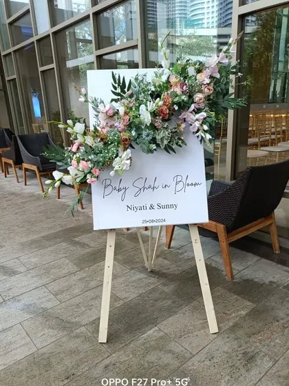 A simple and elegant welcome easel for the "Baby Shah in Bloom" shower. The sign is adorned with a beautiful arrangement of pastel flowers and greenery, setting a sophisticated tone for the event.