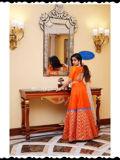 Chaitra Vasudevan seen from the back, showing off the fit and design of the orange lehenga set. The simple blouse back complements the rich texture of the brocade skirt.