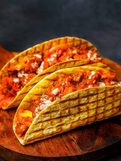A pair of grilled tacos filled with spicy paneer or chicken, served on a wooden board. The dark, moody lighting highlights the texture of the grilled tortilla and the creamy filling.