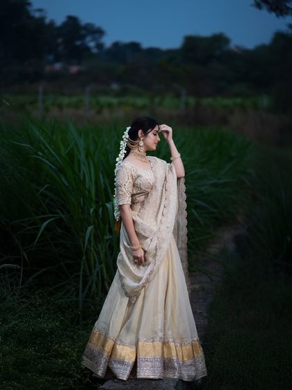 A wide-angle shot capturing the serene beauty of the model in a traditional half saree against the backdrop of a sugarcane field at dusk.
