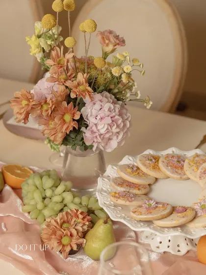 A close-up of the thoughtful details on an anniversary brunch table. A plate of floral-pressed cookies sits beside a small vase of craspedia and daisies, with fresh fruit adding a touch of color.