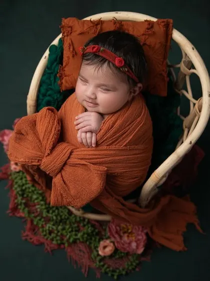 A cozy autumn theme, with a baby swaddled in a rust colored wrap and sleeping in a wicker chair prop against a deep green background.