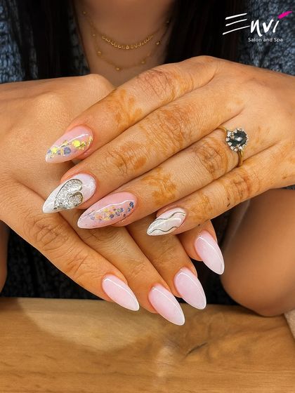 A beautiful example of modern nail art on long, almond-shaped nails. This set features a soft pink base with delicate silver glitter hearts and elegant white swirls.