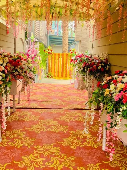 An indoor walkway decorated for a wedding celebration. The path is lined with large floral arrangements in white planters, leading guests into the main event area.