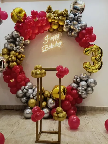 A bold and stylish third birthday decoration. This circular balloon arrangement uses red, silver, and gold chrome balloons, a 'Happy Birthday' neon sign, and a large number '3' foil balloon.
