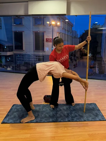 Personal attention is at the heart of what we do. Here, an instructor provides hands-on assistance during a Stick Yoga session, ensuring proper alignment and helping the student safely deepen their backbend in Chakrasana (Wheel Pose).